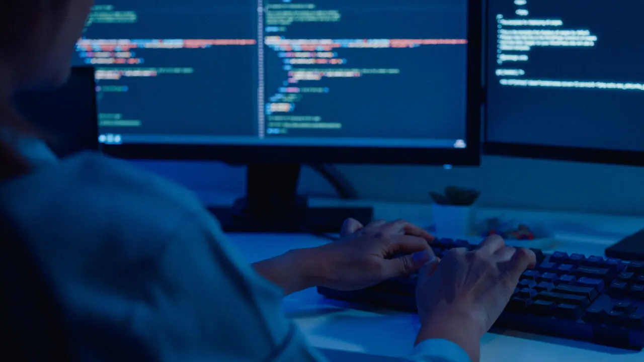 Closeup of young Asian woman API developers using computer to write code sitting at desk with multiple screens work remotely in home at night.