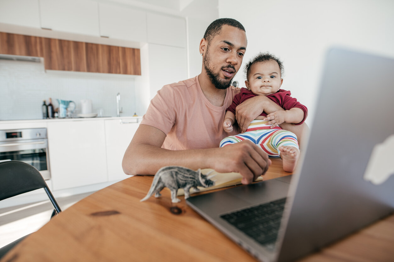 Working dad holds baby while reading his laptop